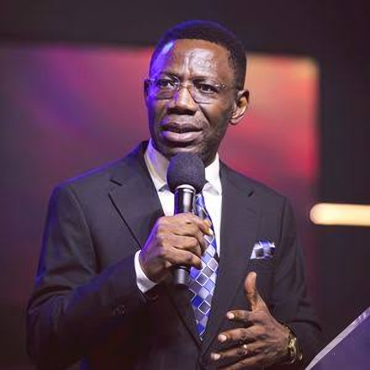 Rev. Dr. Sam Oye preaching on stage, microphone in hand, in a dark suit and blue patterned tie